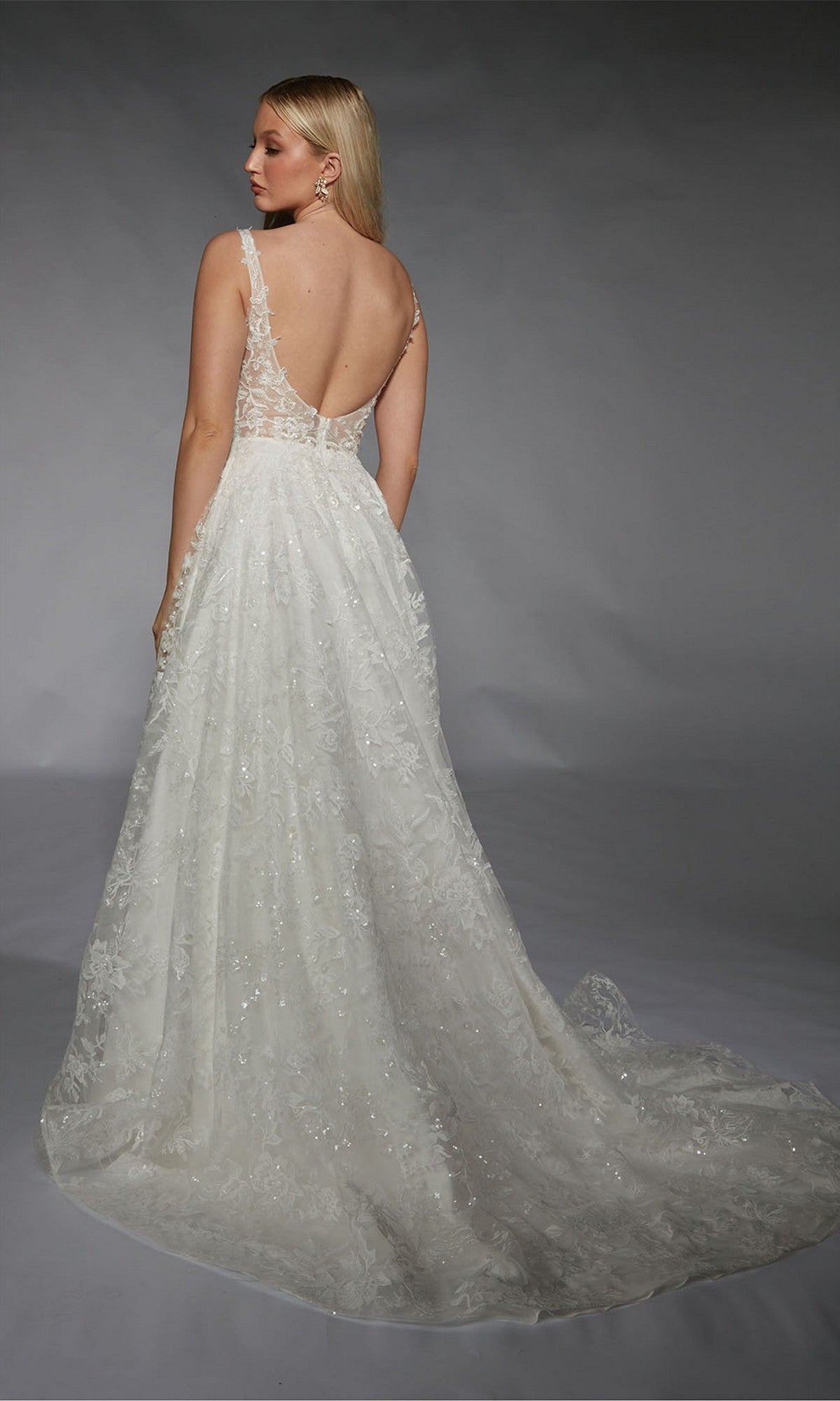 A woman in the Alyce 7123, a sleeveless white A-line wedding gown with lace embellishments, deep V-back, and long train, stands facing away; the dress flows gracefully against a plain background.
